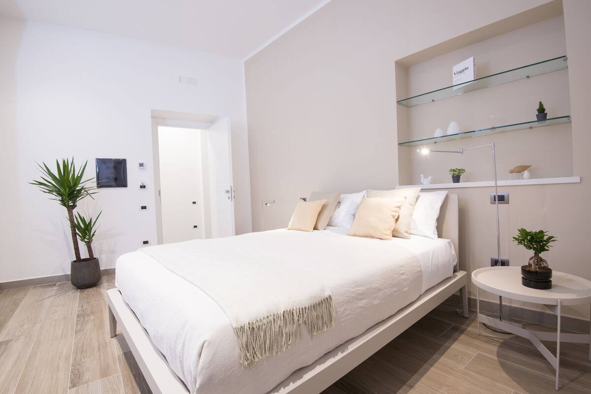 Une chambre à coucher moderne avec un grand lit, une table de chevet agrémentée d’une plante et une étagère en verre placée au-dessus du lit.