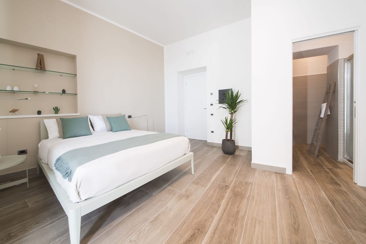 Une chambre moderne avec un grand lit, une table de chevet, une étagère en verre ornée d’objets décoratifs et une plante en pot.