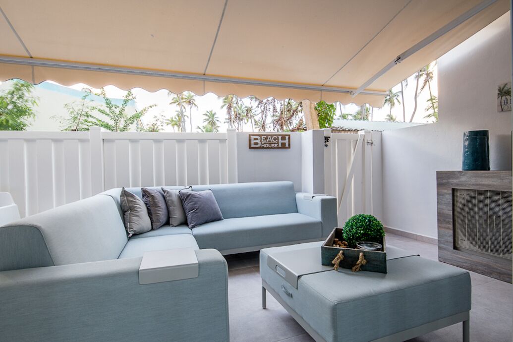 A modern living room with a light blue sofa, a matching ottoman, and a small table with decorative items. A sign reading "BEACH HOUSE" is visible on the wall.