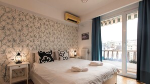 Une chambre avec un grand lit, une table de chevet, un climatiseur fixé au mur, une fenêtre avec des rideaux et une vue sur les bâtiments.
