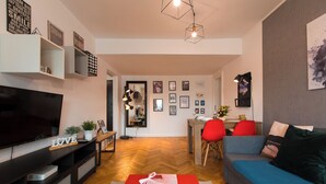 Un salon moderne doté d’un téléviseur à écran plat, d’un canapé, d’un coin repas avec une chaise rouge et d’une cuisine visible à travers une porte ouverte.