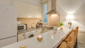 Een moderne keuken met witte kasten, een ingebouwde oven, een magnetron, een spoelbak en een fornuis. Er staat een wasmachine en een klein eiland met een kaars en een plantje.