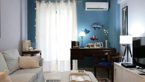 Ein Wohnzimmer mit Sofa, Hocker und einem Schreibtisch mit Computer. Es gibt ein großes Fenster mit Vorhängen, eine Pendelleuchte und eine Wandklimaanlage.