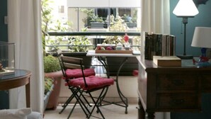 Ein Zimmer mit Balkon, einem Esstisch mit Stühlen, einer Lampe und einem Fenster mit Vorhängen.