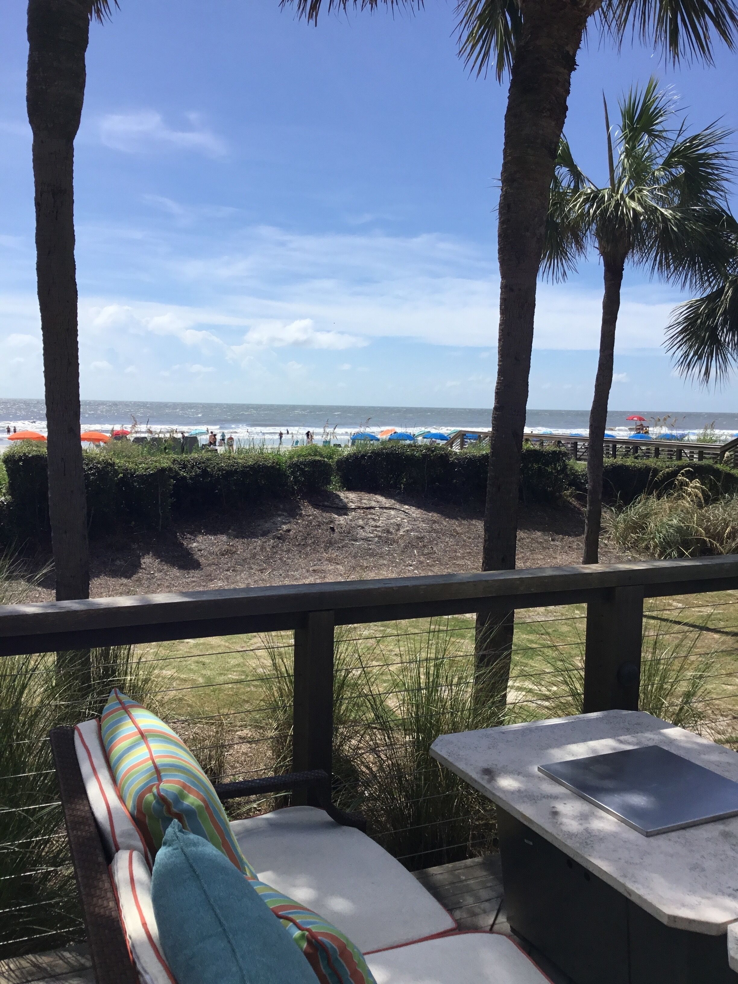 A beach view from a covered deck with a lounge chair and a table.