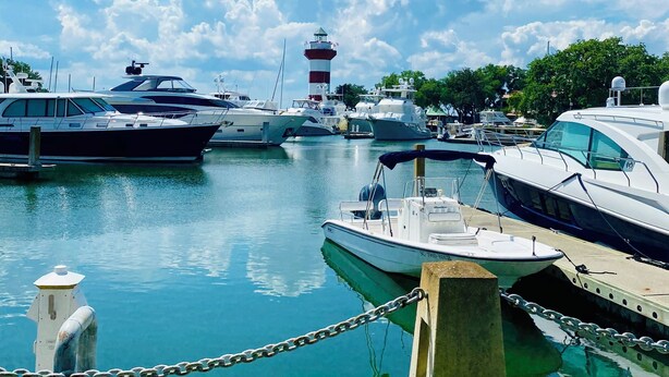 A marina with various boats docked, a lighthouse in the background, and a grassy area in the foreground.