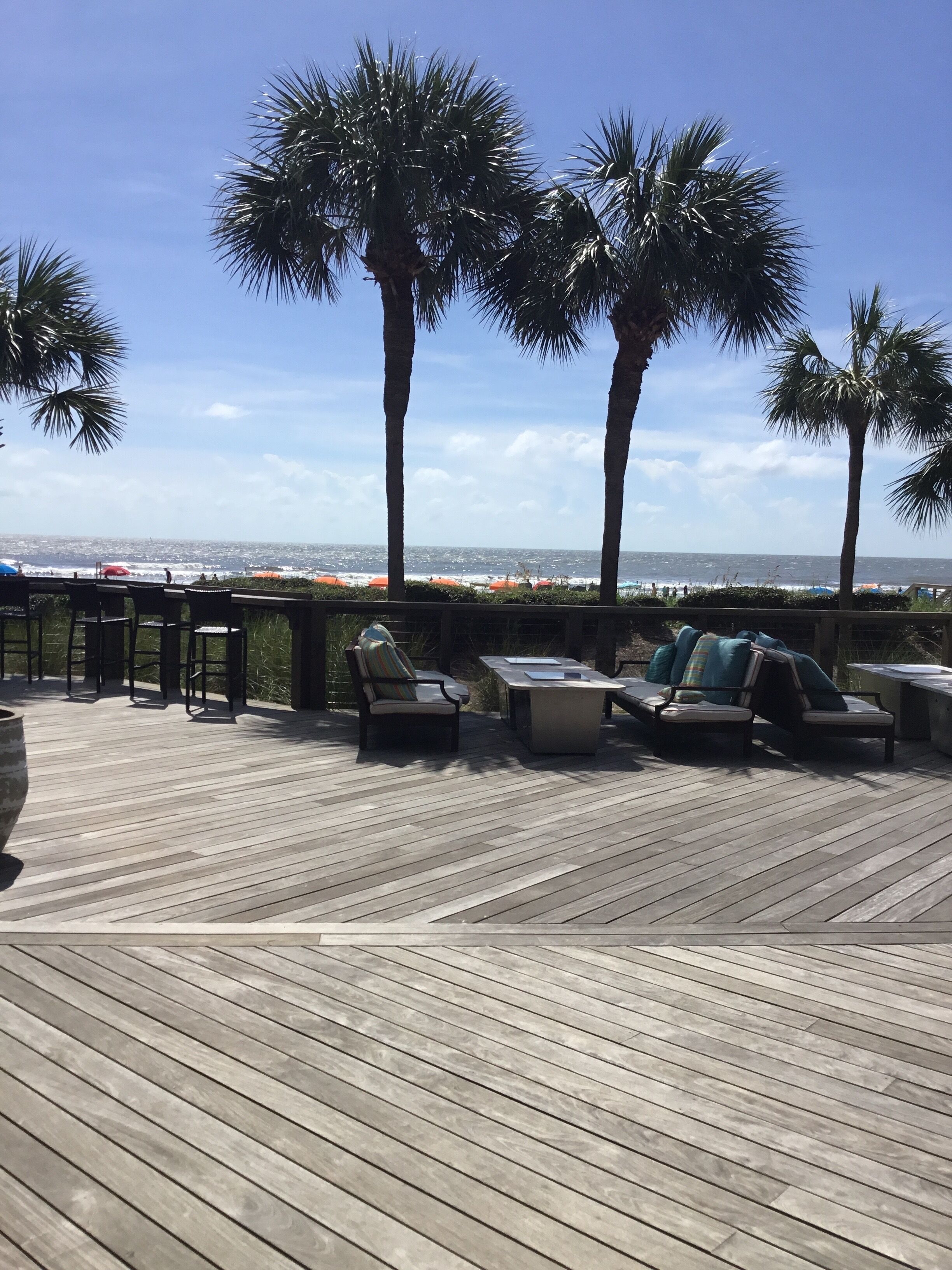 A wooden deck with outdoor seating, palm trees, and a view of the ocean.