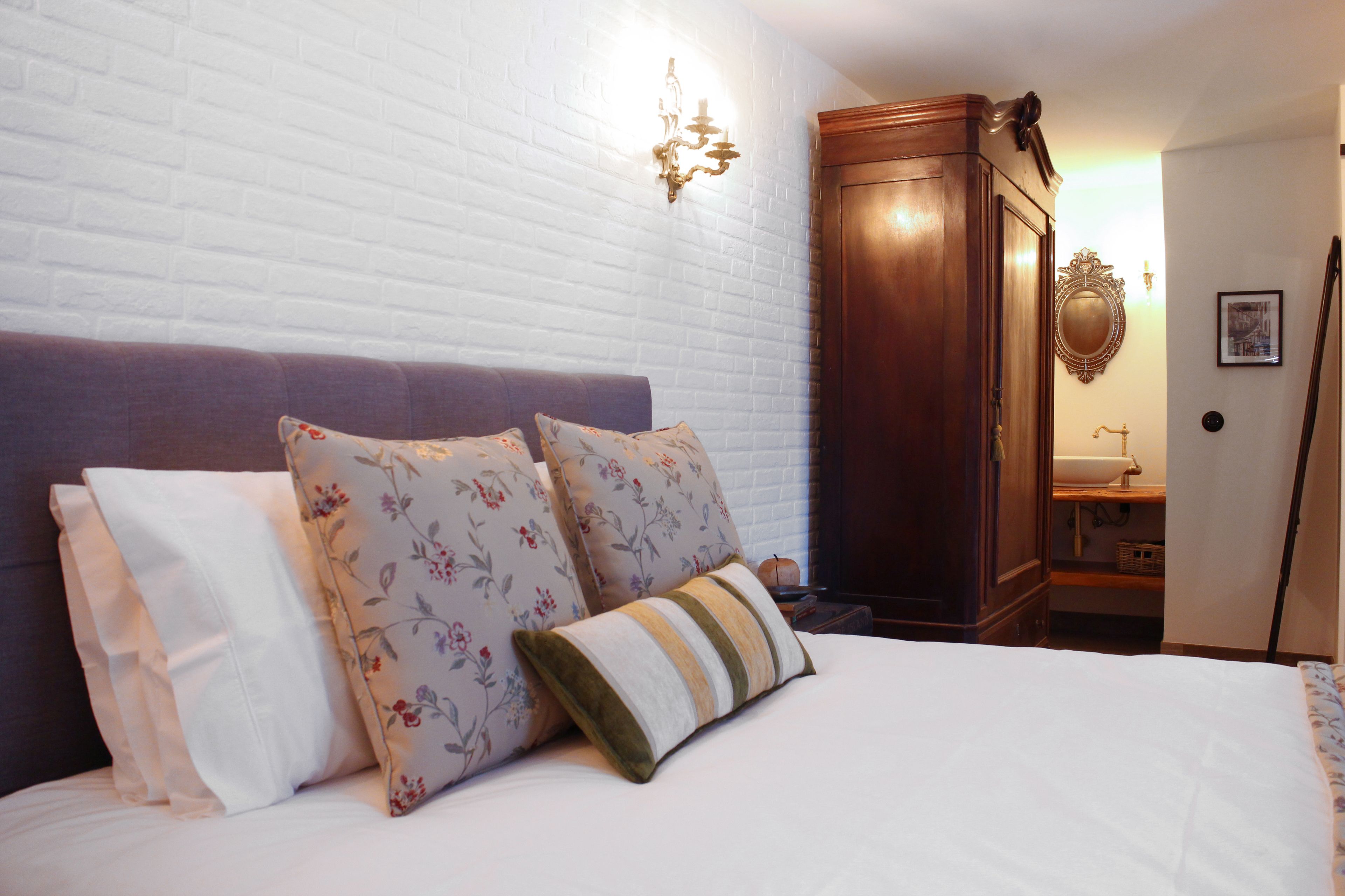 A neatly made bed with floral and striped pillows, a wooden wardrobe, and a wall-mounted mirror.