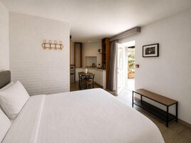 Habitación de hotel con una cama grande, una mesita pequeña, un banco, una kitchenette y un baño visible a través de una puerta abierta.