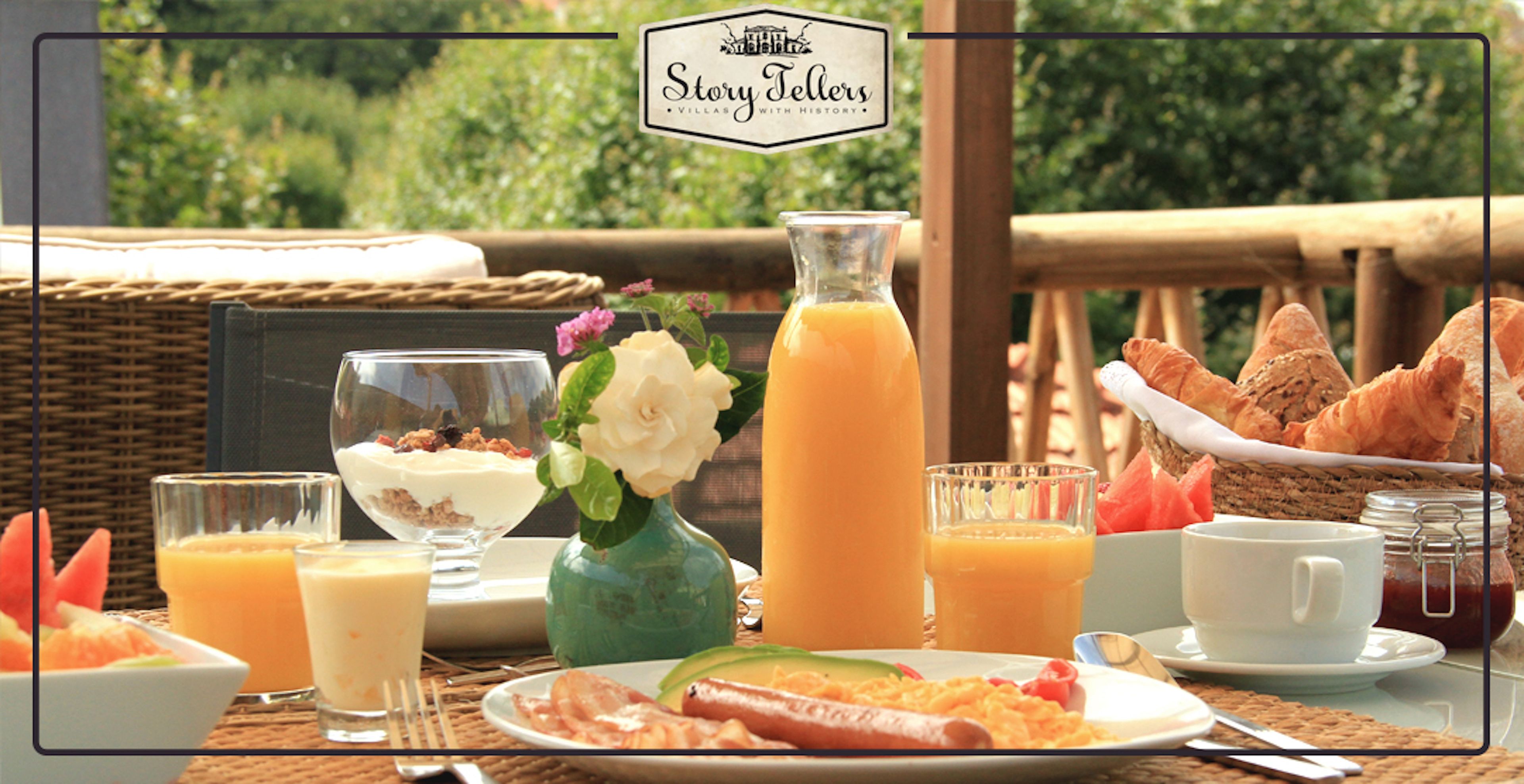 A breakfast setting with a wicker table, a vase with a flower, and a basket of croissants.