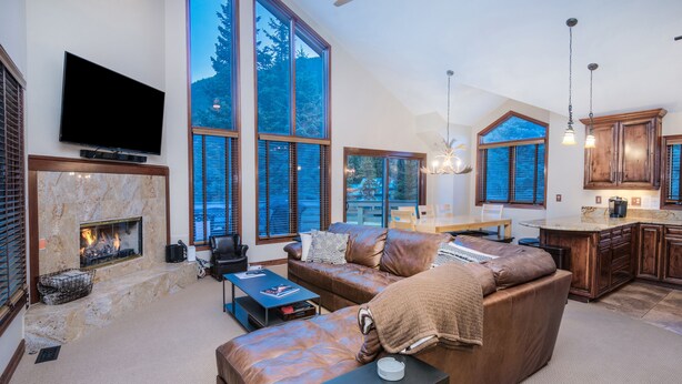 A spacious living room with a fireplace, large windows, a television, and a kitchen area. - Nice Home in Great Vail Location! Hot Tub, Free Shuttle Access!! (Vail)
