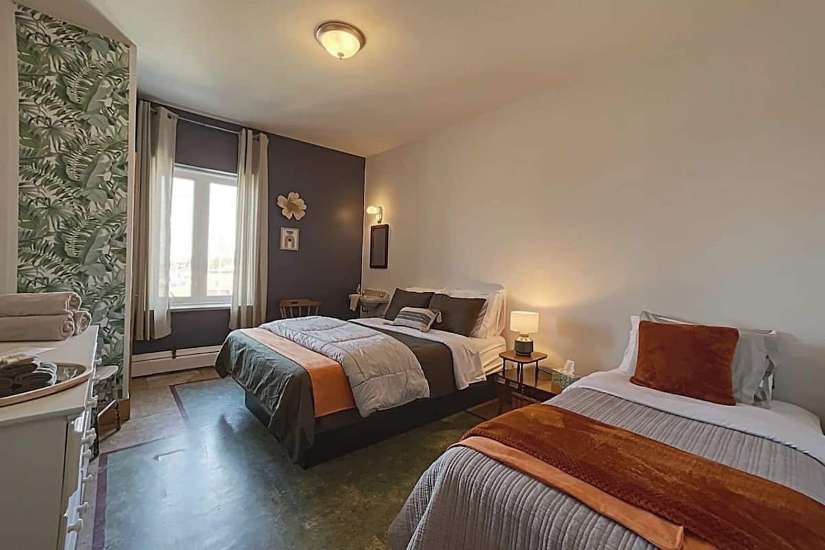 Ein Hotelzimmer mit zwei Betten, einem Schreibtisch, einem Stuhl, einem Fenster mit Vorhängen, einer Wand mit tropischem Blattmuster und einer Deckenleuchte.