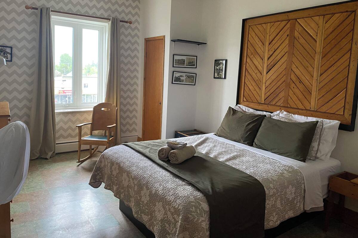 Ein Schlafzimmer mit einem Bett, einem hölzernen Kopfteil, einem Sessel, einem Fenster mit Vorhängen und einer Wand mit gerahmten Bildern.