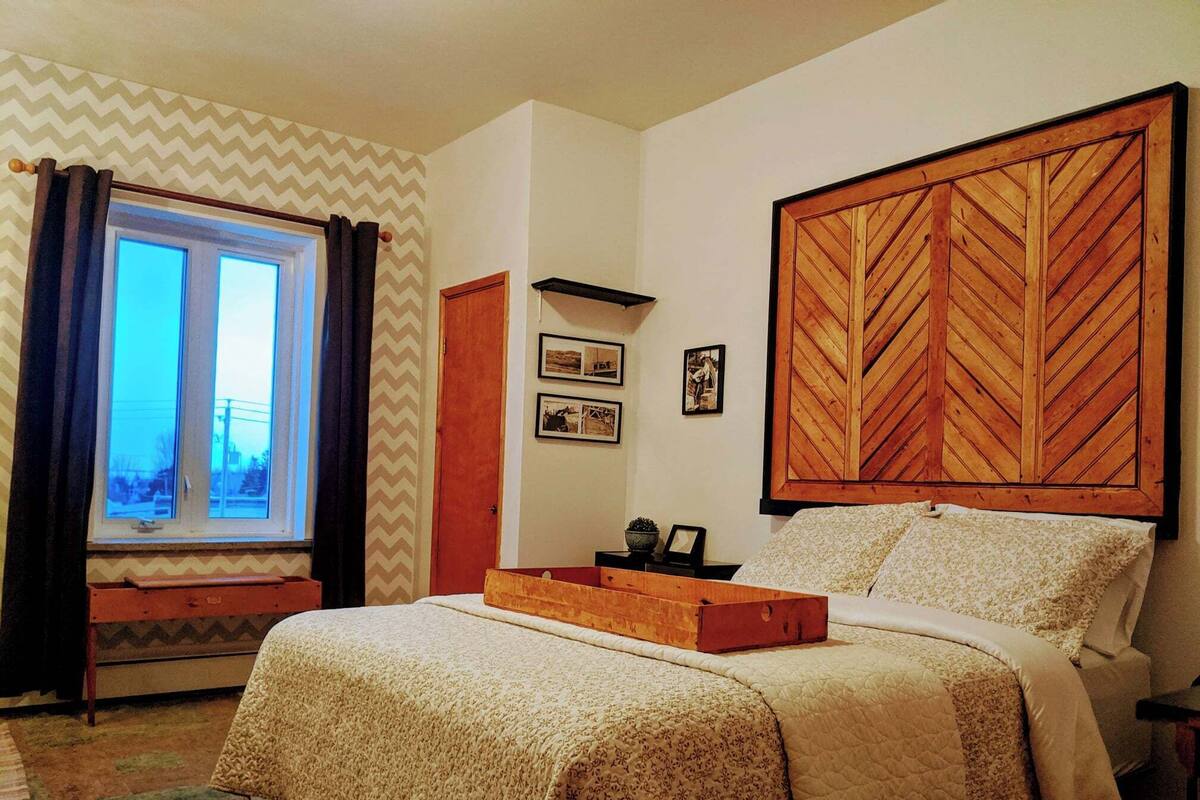 Ein Schlafzimmer mit Bett, Fenster mit Vorhängen, einem Holzkopfende und einem an der Wand befestigten Regal mit Bildern.