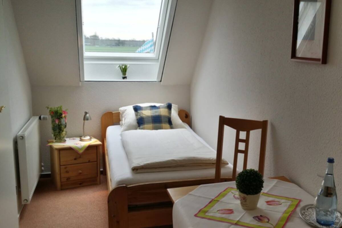 Een kleine, gezellige slaapkamer met een enkel bed, een nachtkastje, een stoel, een klein tafeltje met een plant en een raam met uitzicht.