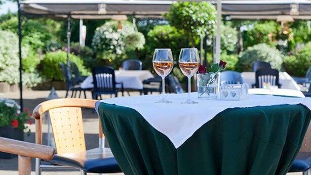 An outdoor dining area with a table covered in a white tablecloth, two wine glasses, and a vase with a single rose. - Landgasthaus Hotel Zur Alten Linde (Hamersen)
