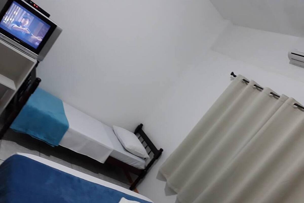 Une chambre d’hôtel avec deux lits, l’un recouvert d’une couvre-lit bleue et l’autre d’une couvre-lit blanche. Il y a un téléviseur posé sur un meuble à gauche de la pièce, et une fenêtre avec des rideaux blancs.
