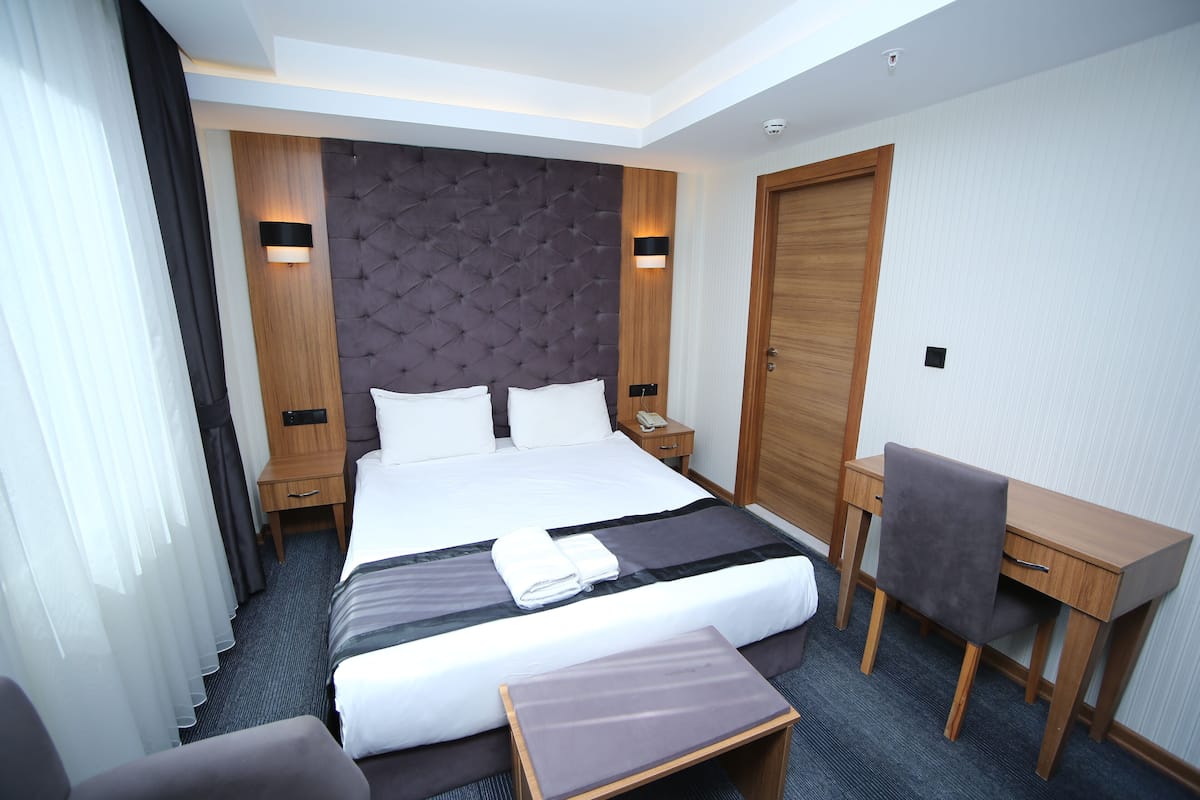 Habitación de hotel con una cama grande, un escritorio y una silla. Hay dos mesitas de noche, una con una lámpara y la otra con un teléfono.