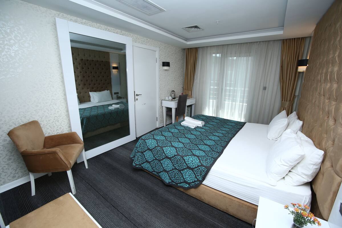 Una habitación de hotel con cama, sillón, escritorio, espejo y ventana con cortinas.