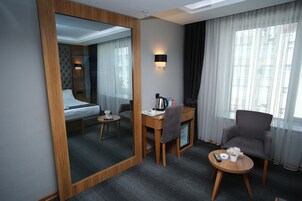 A hotel room with a bed, a desk with a chair, a small table with a remote control, a coffee machine, and a decorative plant.