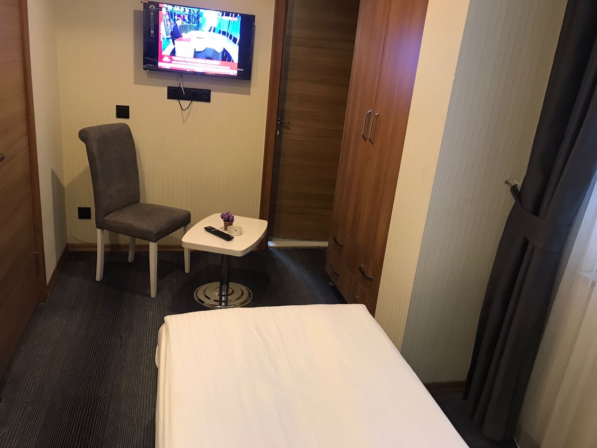 Ein Hotelzimmer mit einem Bett, einem kleinen Tisch mit Fernbedienung, einem Sessel und einem an der Wand befestigten Fernseher.