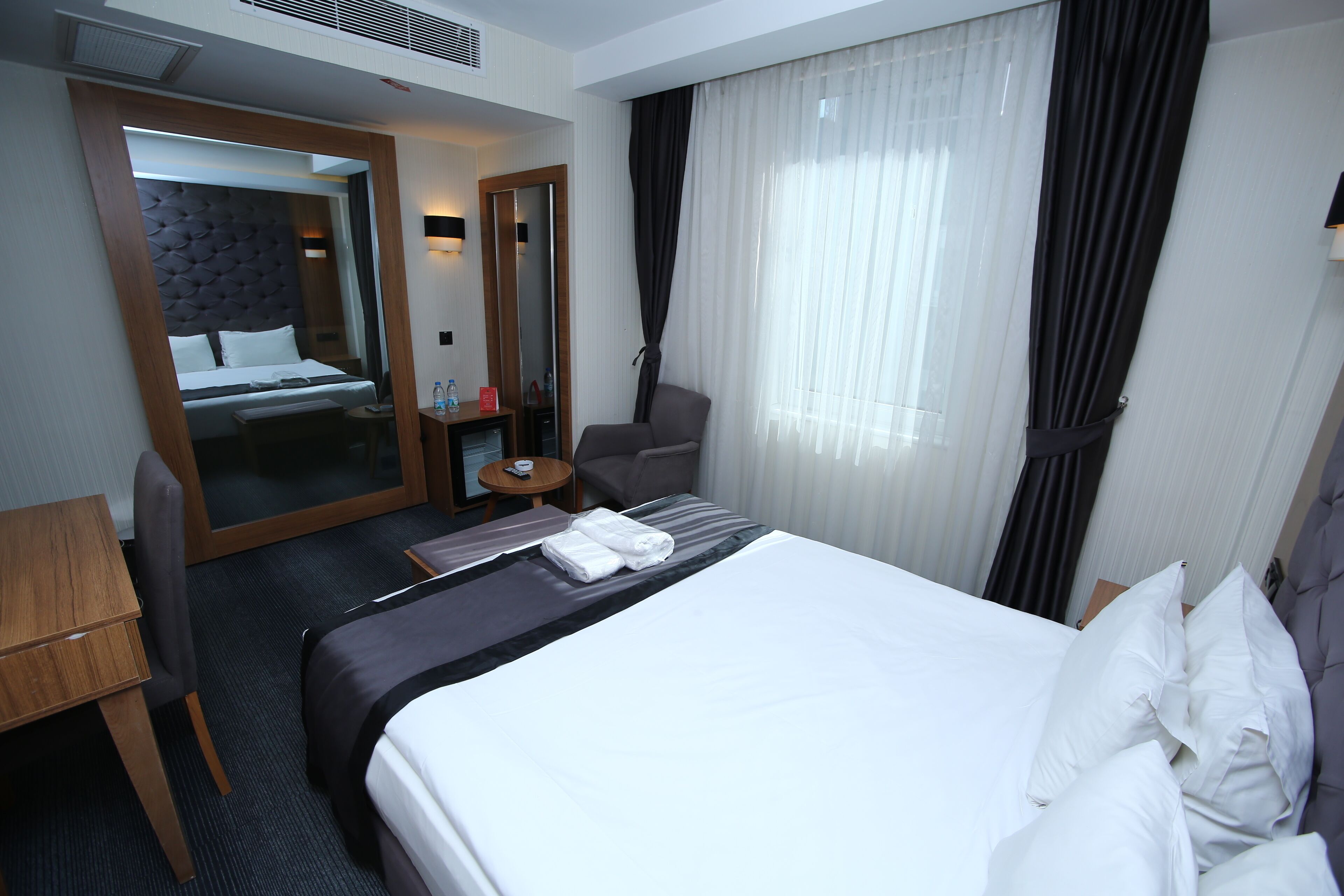 A hotel room with a large bed, a desk, a chair, a mirror, and a small table with a bottle and a red object.