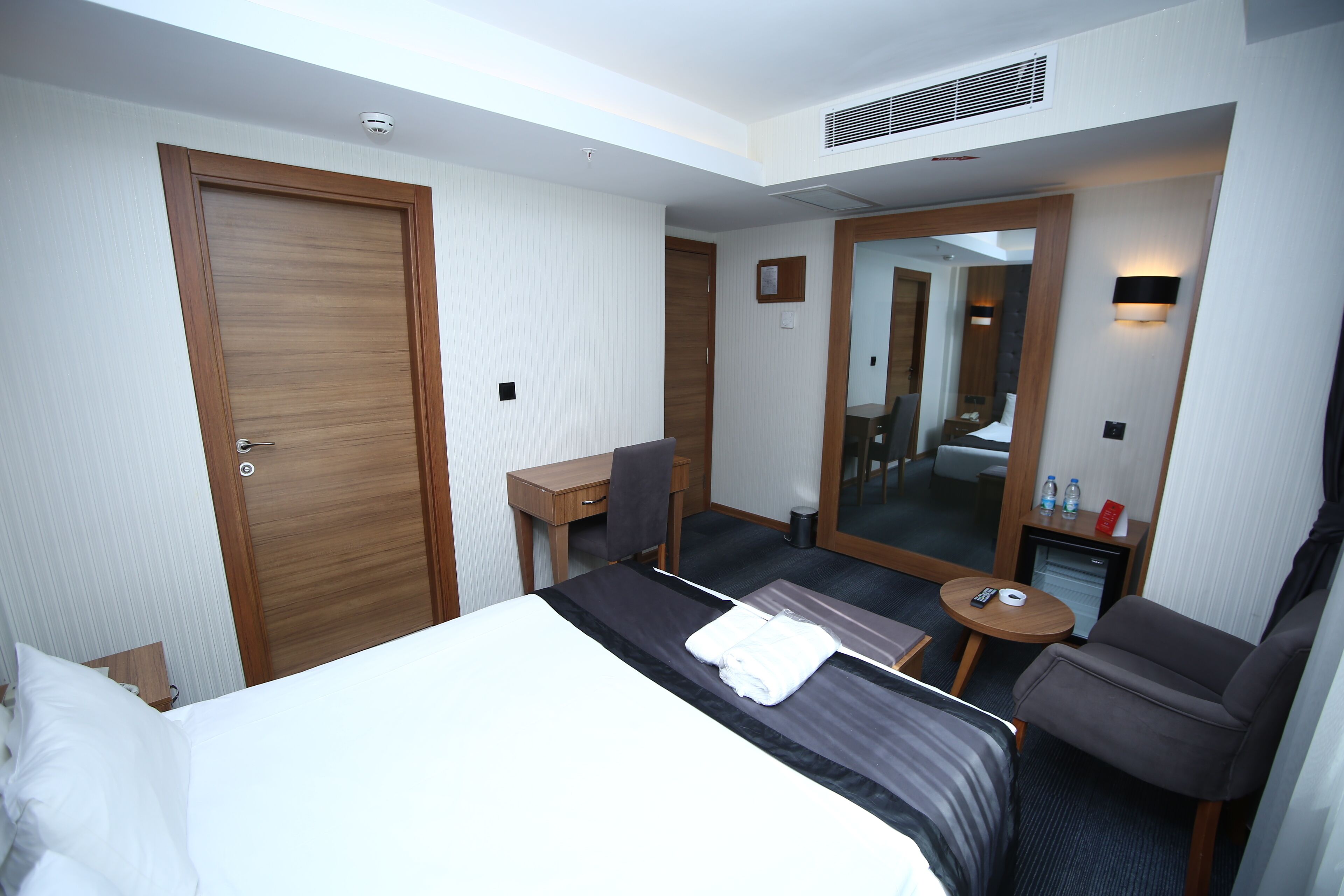 A hotel room with a bed, a desk, a chair, a small table, and a mirror.