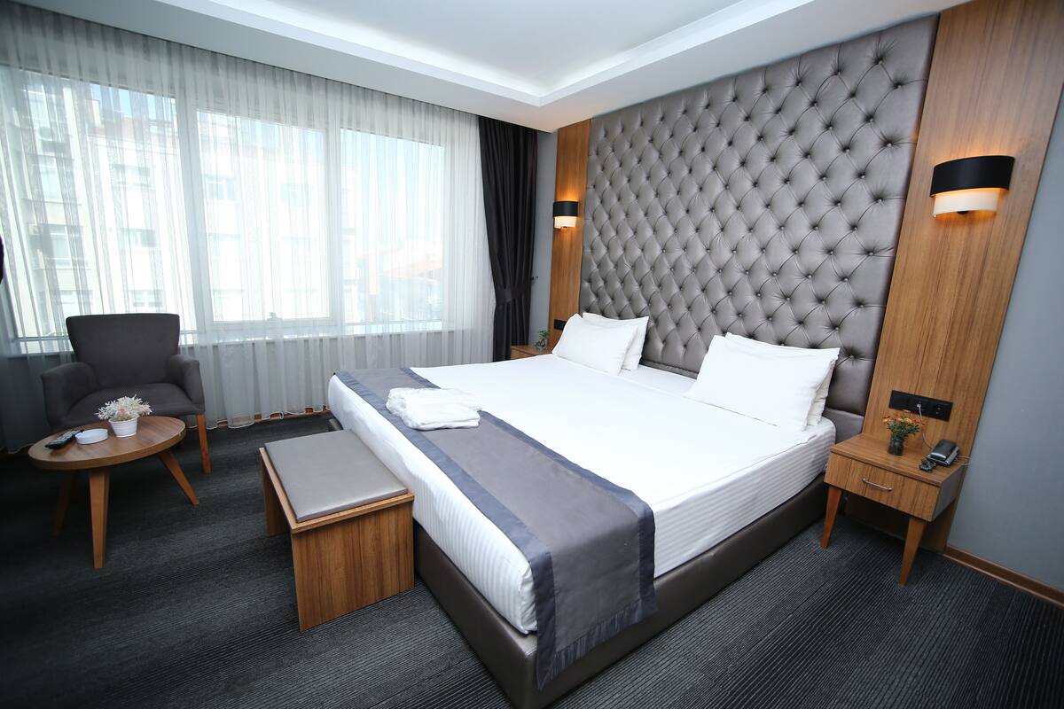 Habitación de hotel con una cama grande, una mesita de noche, una silla, una pequeña mesa con un control remoto y una ventana con cortinas.