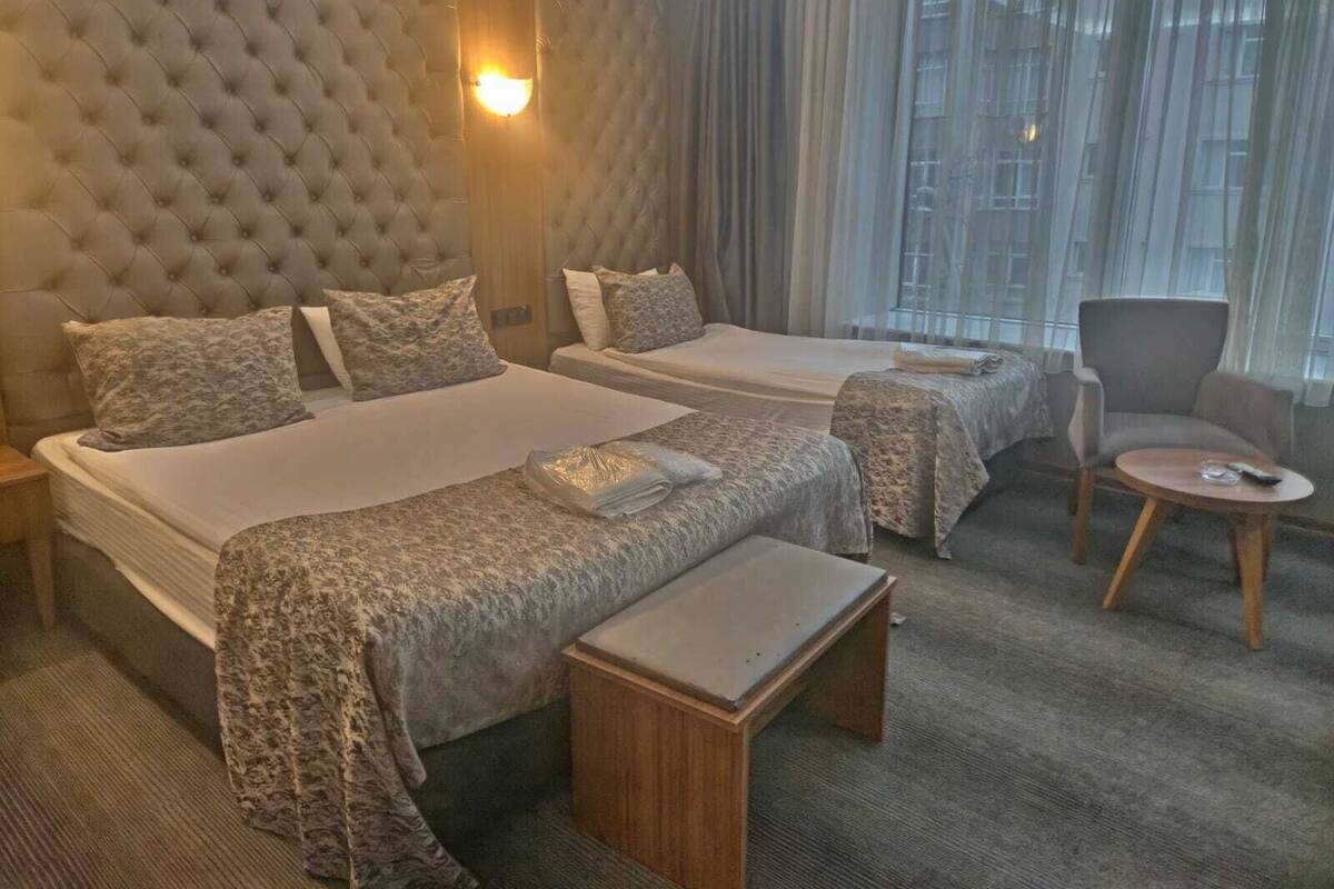 Habitación de hotel con dos camas, una mesita pequeña, una silla y un ventanal grande con cortinas.