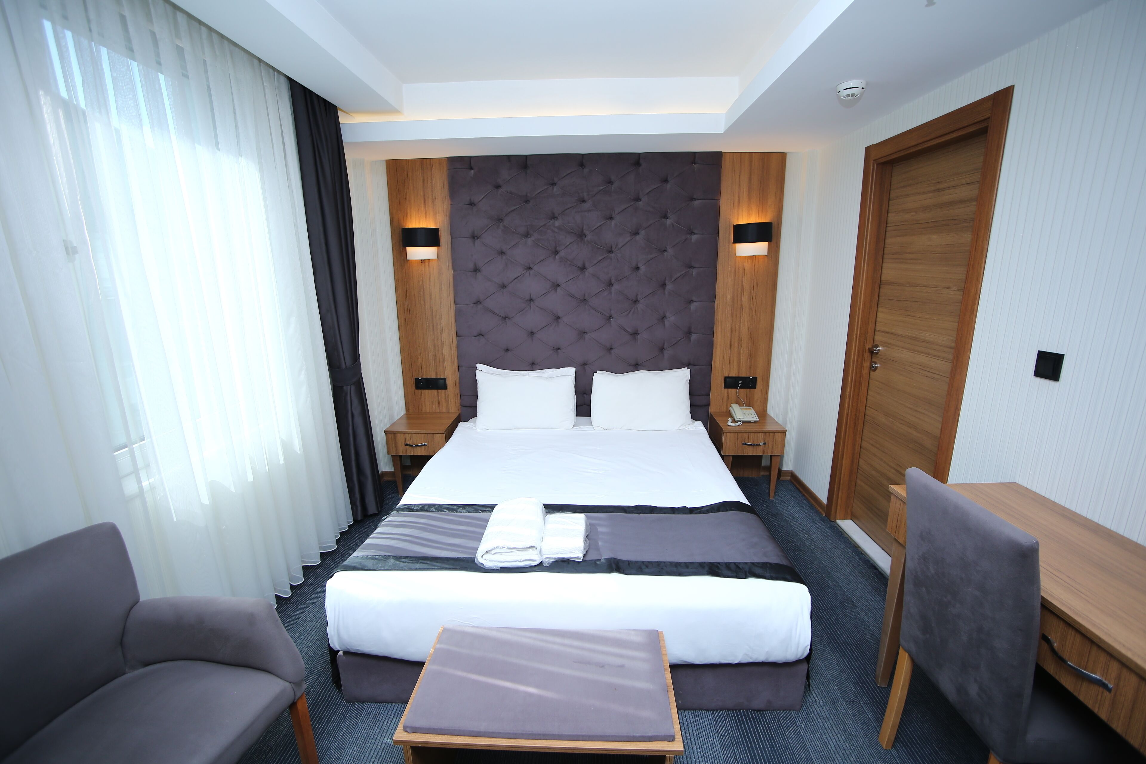Quarto de hotel com cama grande, escrivaninha, cadeira e mesinha.