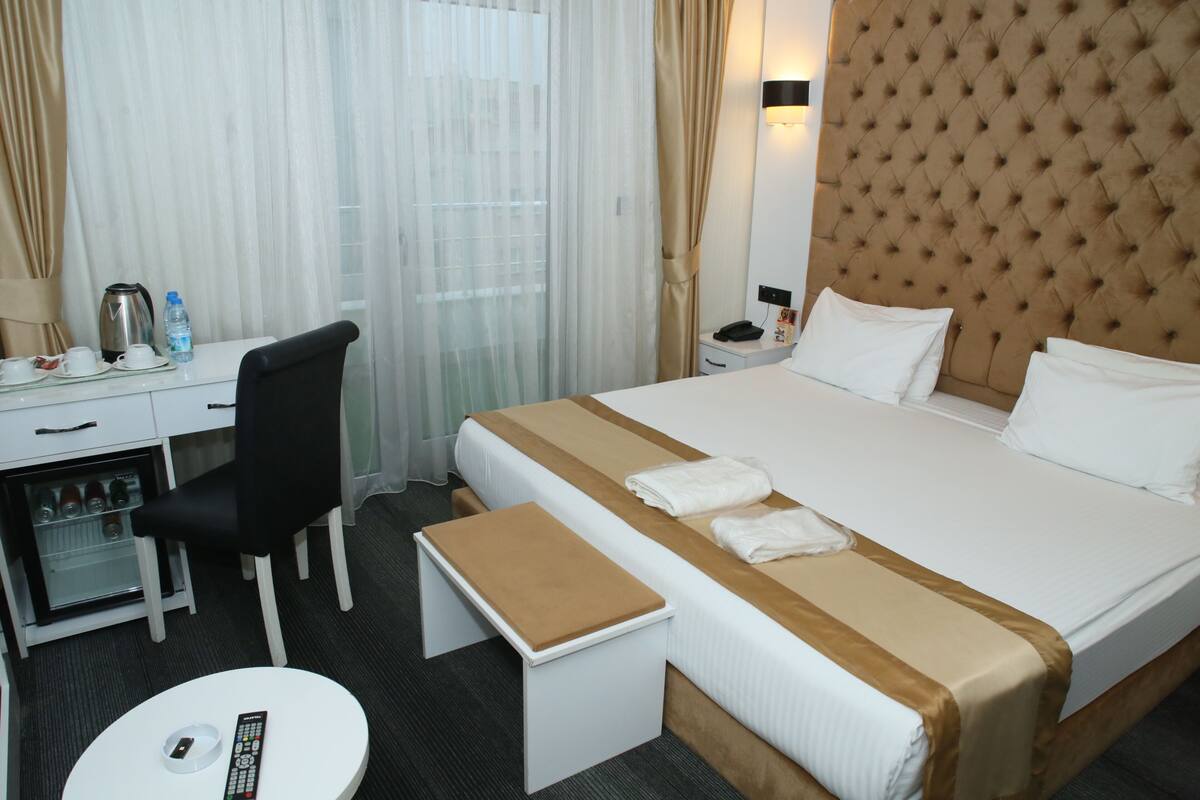 Una habitación de hotel con cama, escritorio, silla, mesita pequeña, mesita de noche y lámpara fijada a la pared.
