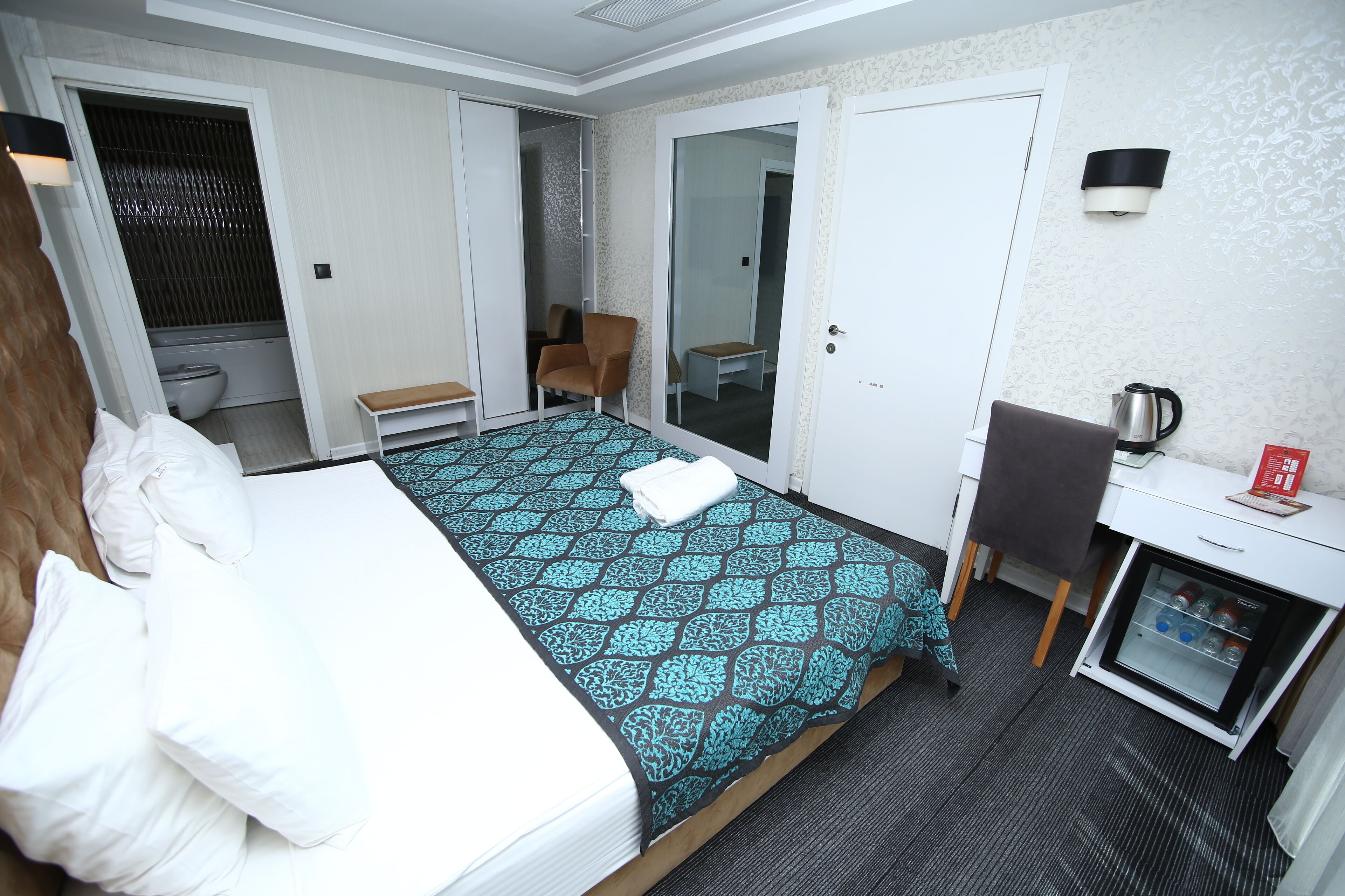 Habitación de hotel con cama, escritorio, silla, refrigerador pequeño, espejo y baño.