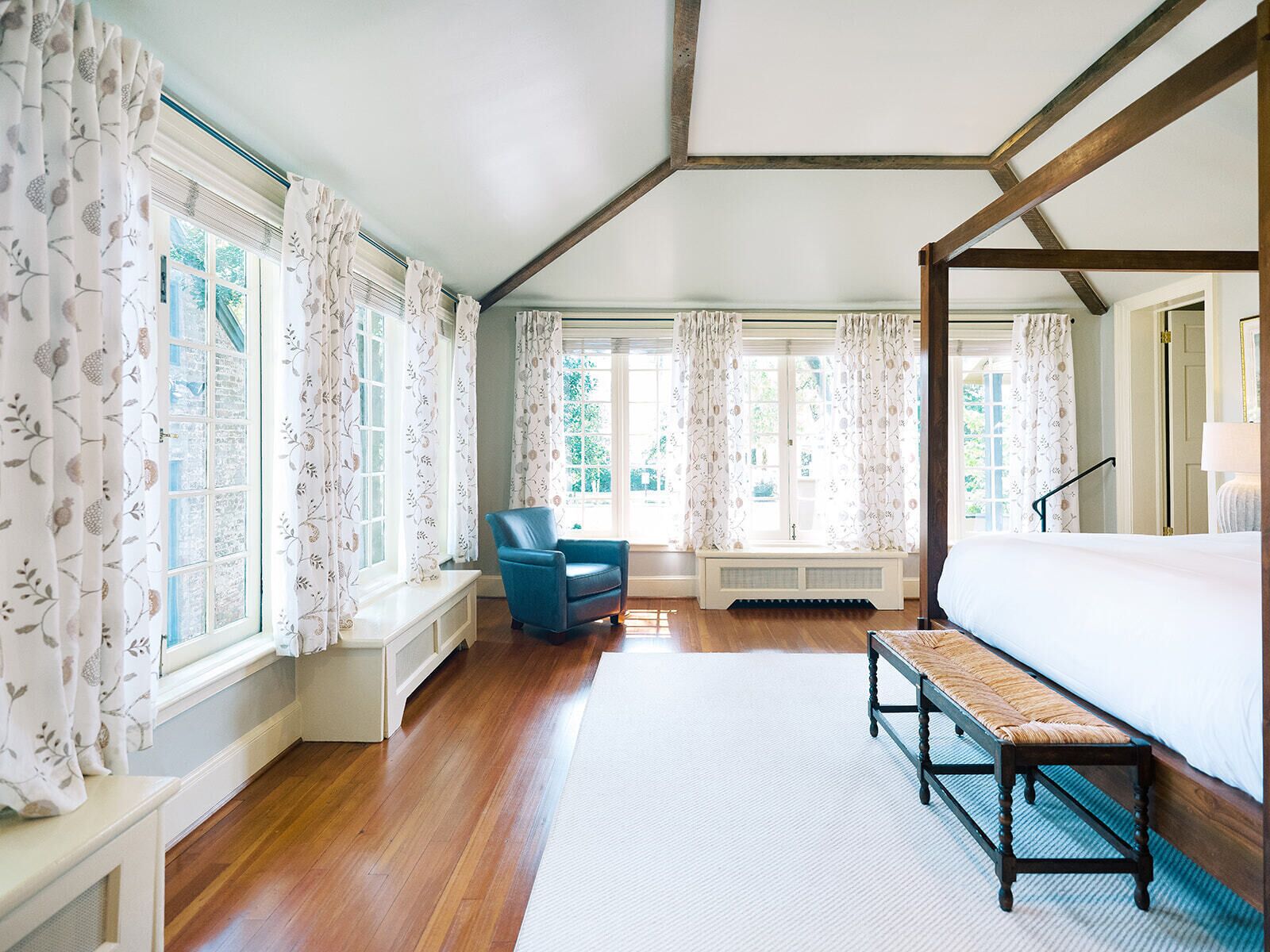 A bedroom with a four-poster bed, a bench, a chair, and large windows with curtains.