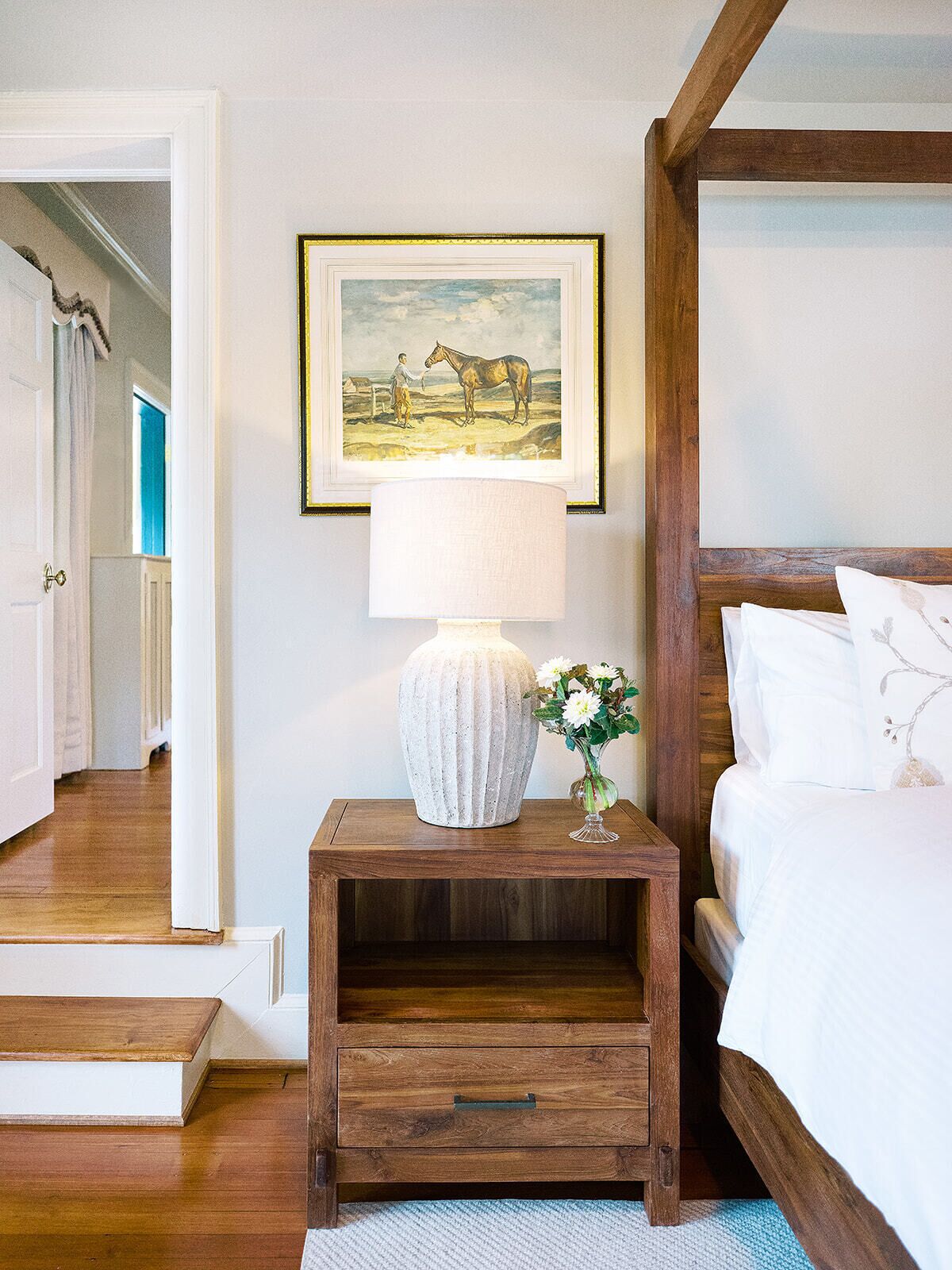 A bedroom with a wooden nightstand, a lamp, a framed picture of a horse, and a bed with white bedding.