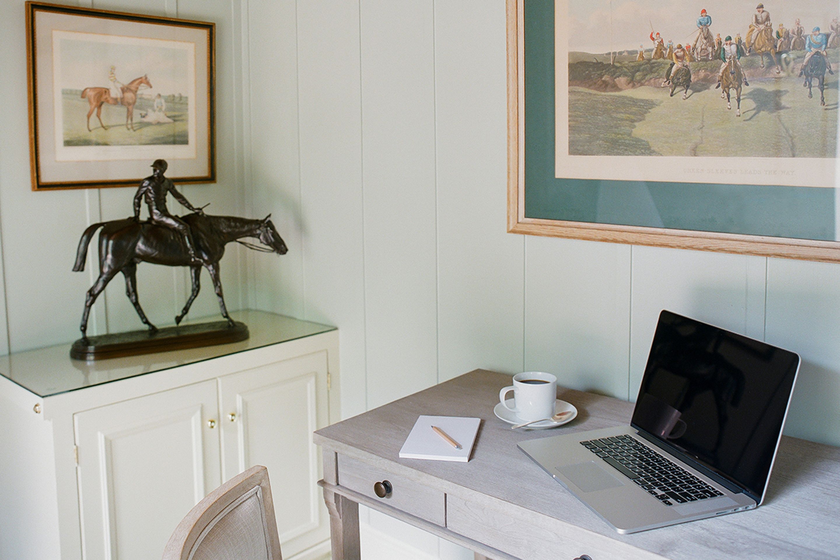 Une pièce comprenant un bureau, un ordinateur portable, une tasse, un tableau représentant un cheval et une chaise.