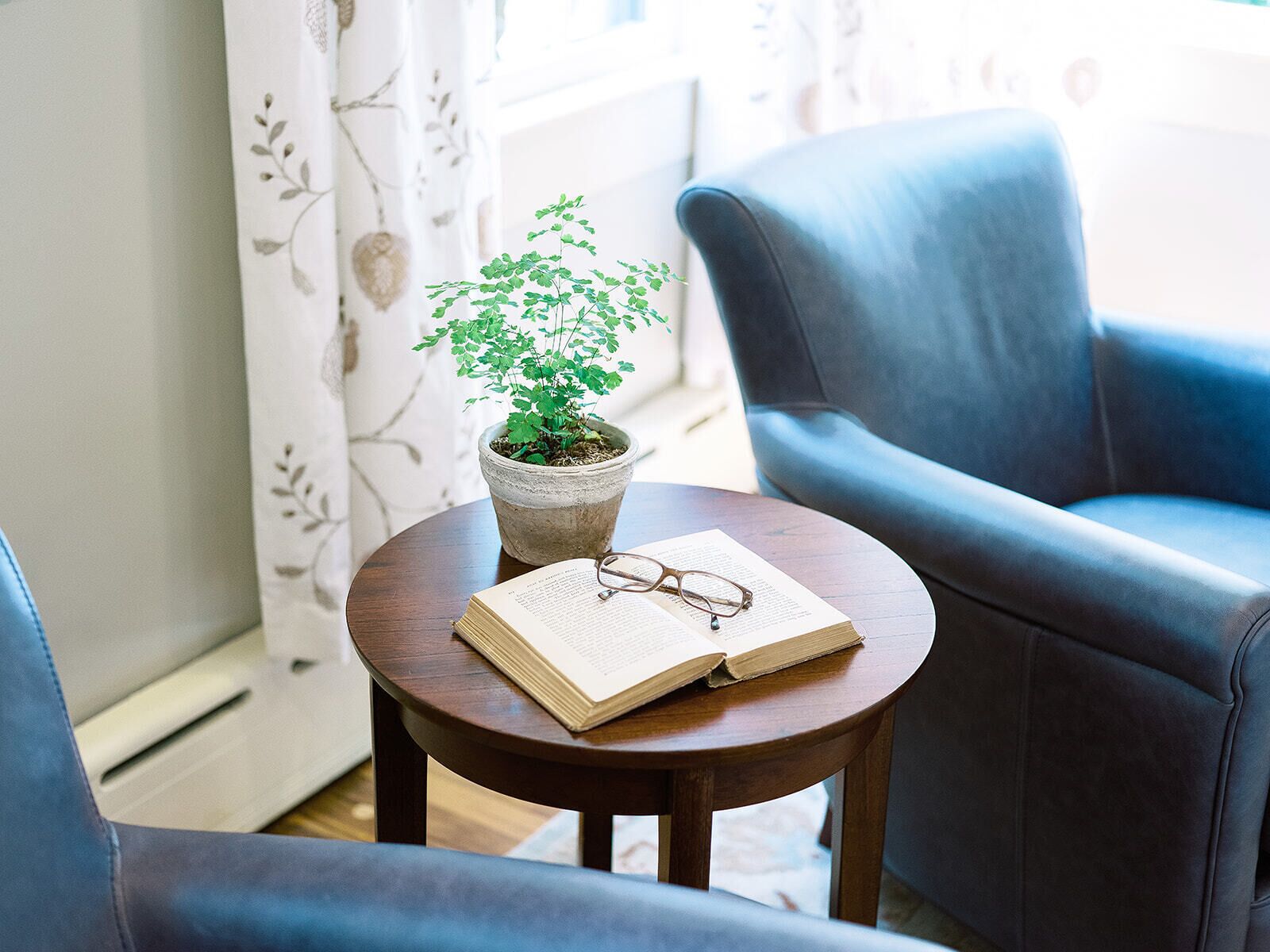 A cozy room with a round wooden table, a potted plant, an open book, and a pair of glasses.