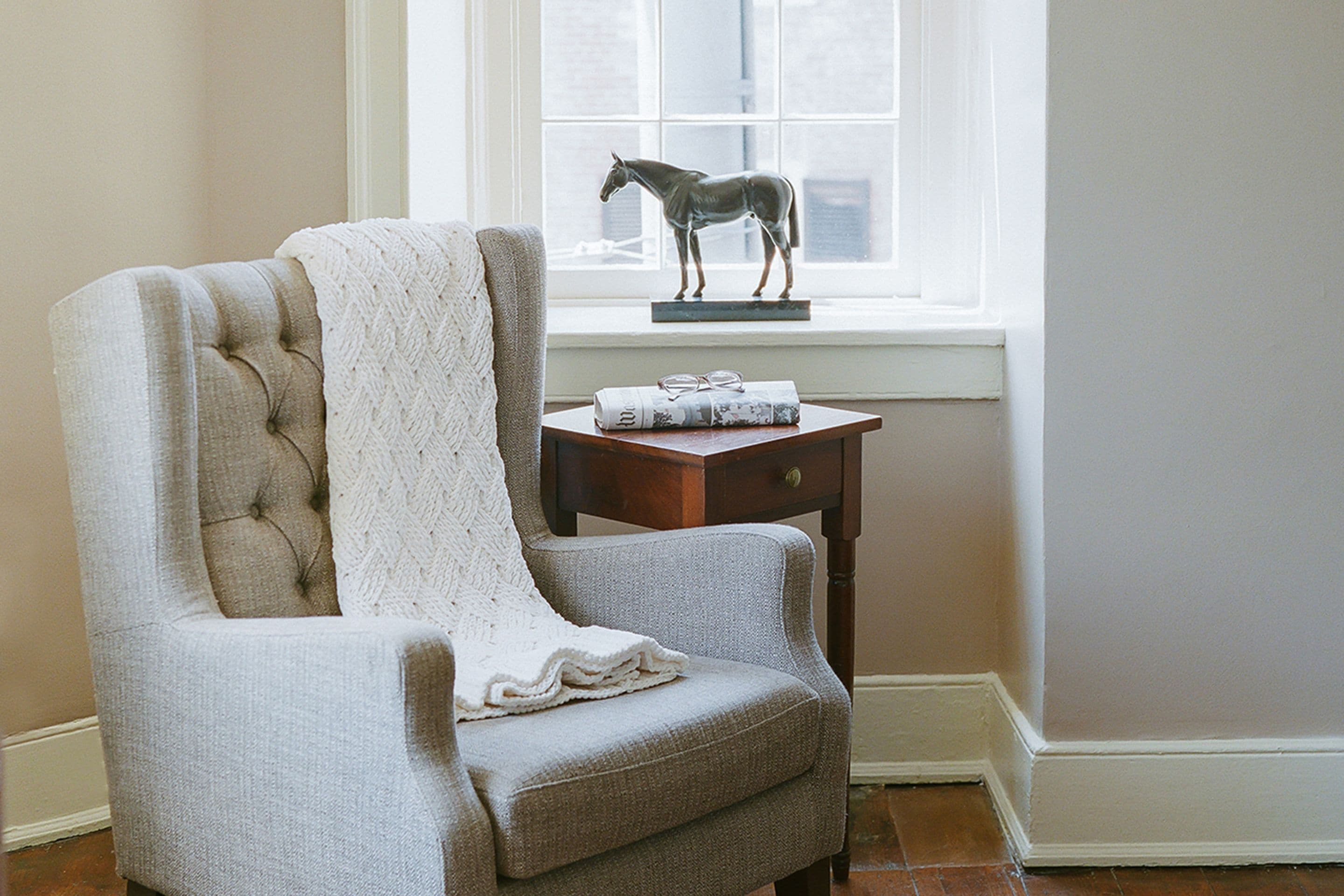 Un sillón con textura, una manta blanca de punto, una mesita de madera con una estatua de un caballo y una ventana con vista a un edificio.