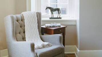 Un sillón con textura, una manta blanca de punto, una mesita de madera con una estatua de un caballo y una ventana con vista a un edificio.