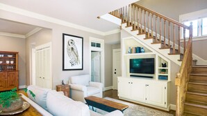A living room with a white sofa, a wooden coffee table, a TV on a white cabinet, and a wooden staircase.