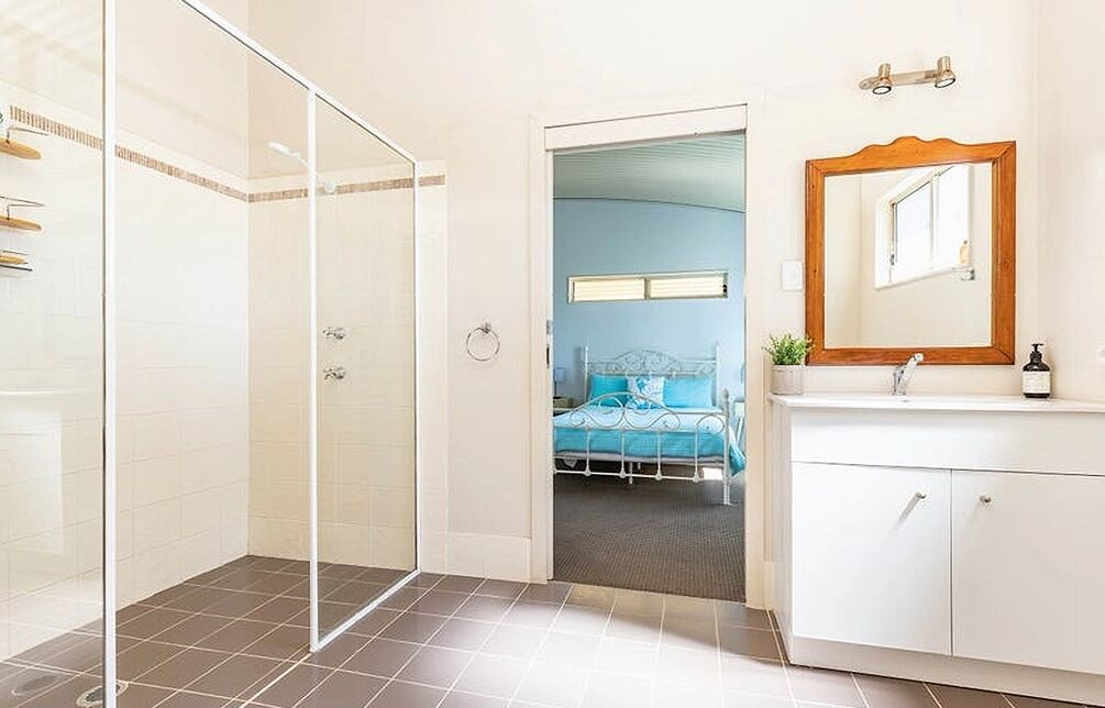 Une salle de bain moderne avec une douche à l’italienne en verre, un meuble-lavabo blanc avec un miroir et un lit dans la pièce adjacente.