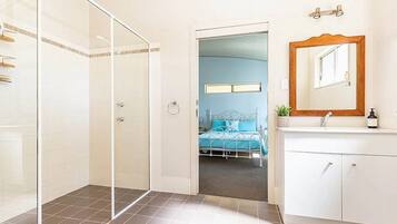 Une salle de bain moderne avec une douche à l’italienne en verre, un meuble-lavabo blanc avec un miroir et un lit dans la pièce adjacente.