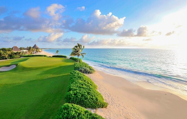 A golf course with a beach view, palm trees, and thatched-roof buildings.