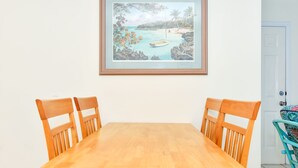 A wooden dining table with four chairs, a framed painting of a beach scene, and a white door in the background.