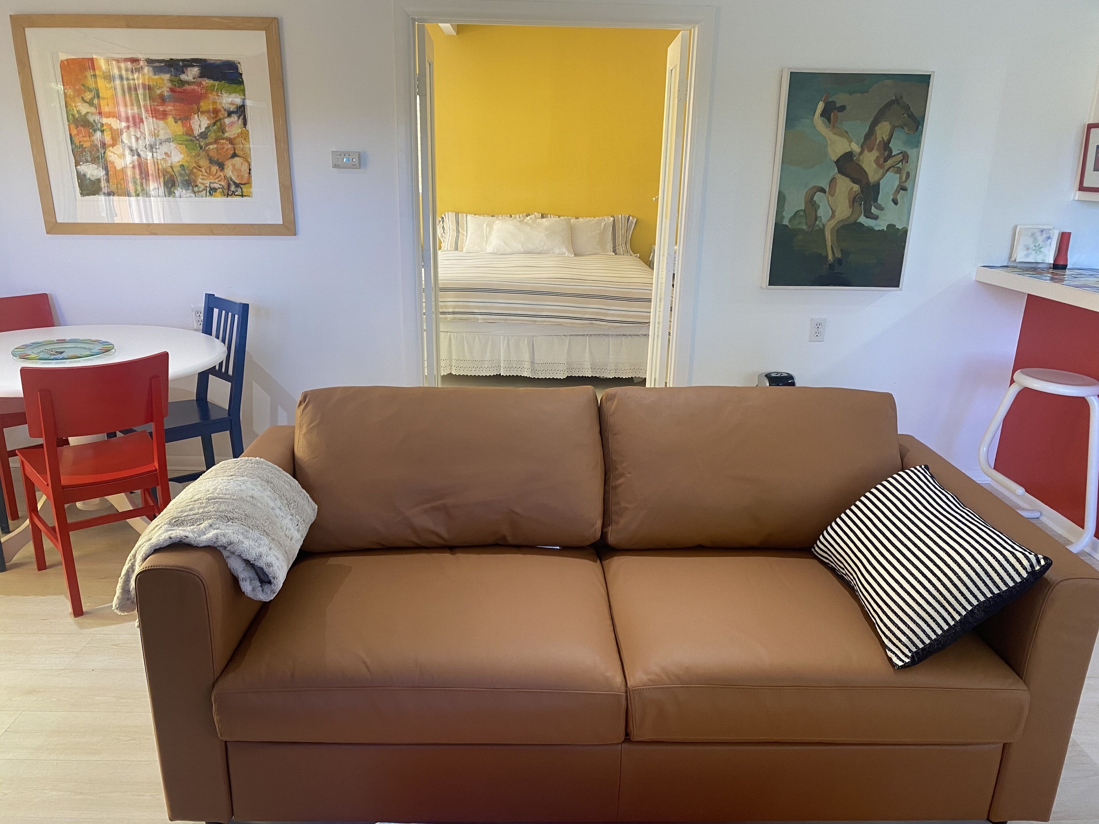 Un salon chaleureux avec un canapé marron, une petite table entourée de chaises, un lit devant un mur jaune et des œuvres d’art accrochées aux murs.