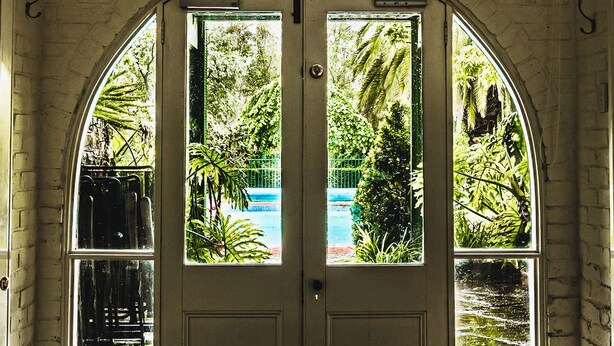 An arched doorway with two glass panels, leading to a courtyard with a fountain and greenery. - Corvah House, Beautiful Corvah House circa 1860, restored and beaming with grandeur and comfort (Castlemaine)