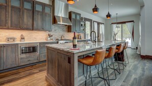 A modern kitchen with dark wooden cabinets, a large island with bar stools, stainless steel appliances, and pendant lighting. - Sunriver retreat; 5bed, 4primarysuite,Theatre rm!! (Sunriver)