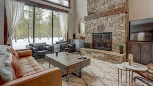 A cozy living room with a stone fireplace, leather sofa, and a large window overlooking a snowy landscape. - Sunriver retreat; 5bed, 4primarysuite,Theatre rm!! (Sunriver)
