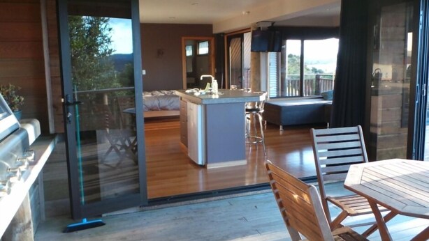 A modern kitchen with a central island, a dining area with a table and chairs, and a sliding glass door leading to a balcony. - Beautiful Waterview Studio (Ngunguru)
