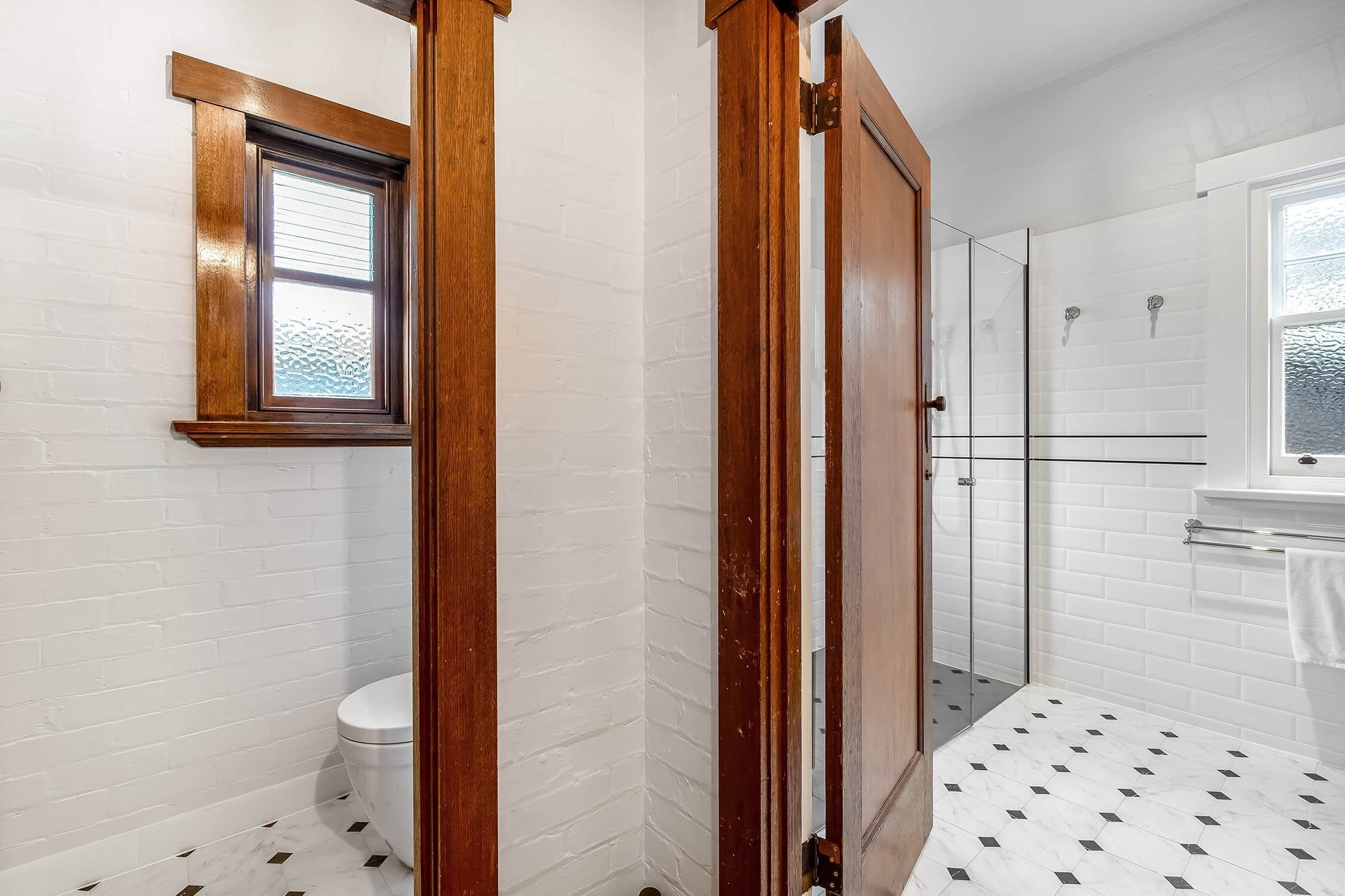 A bathroom with a white brick wall, a toilet, a shower with glass doors, and a window with blinds.