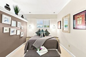 A hotel room with a bed, bedside tables, a window with blinds, wall-mounted artwork, and a potted plant.